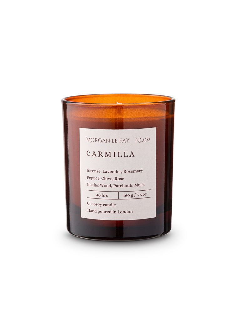 Candle with an amber transparent glass jar and a label that says 'carmilla' on a white background