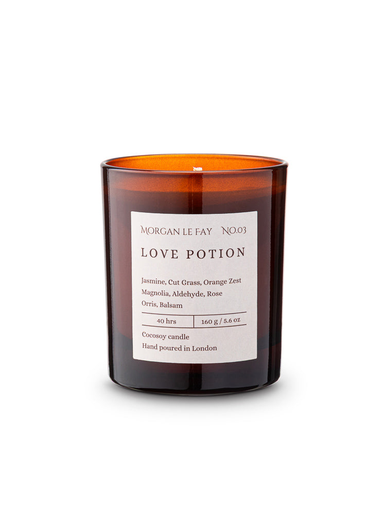 Candle with an amber transparent glass jar and a label that says 'love potion' on a white background