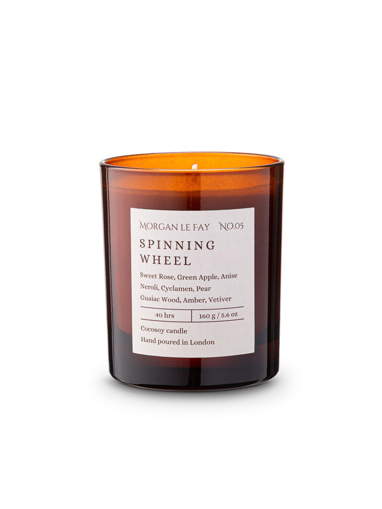 Candle with an amber transparent glass jar and a label that says 'spinning wheel' on a white background