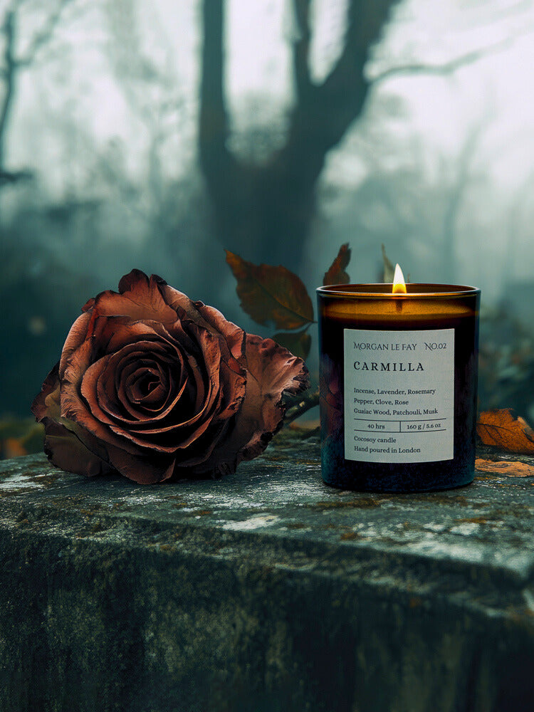 Candle labeled 'Carmilla' next to a dried rose on a stone surface with a forest background