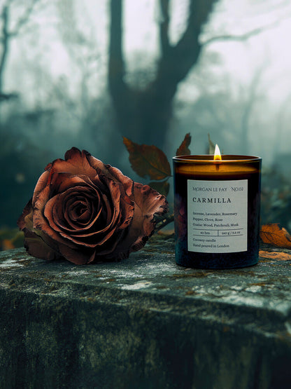 Candle labeled 'Carmilla' next to a dried rose on a stone surface with a forest background