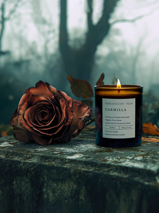 Candle labeled 'Carmilla' next to a dried rose on a stone surface with a forest background
