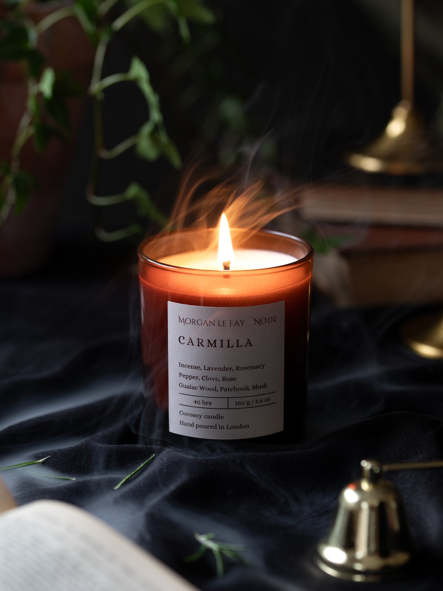 Candle labeled 'Carmilla' burning on a dark fabric surface with plants and a bell in the background.