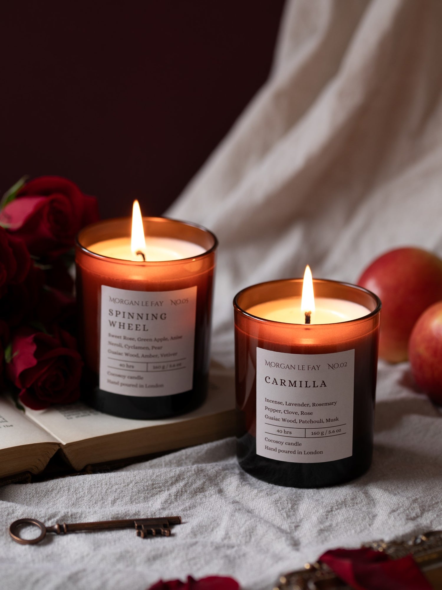Two lit candles with labels on a surface with apples and a book