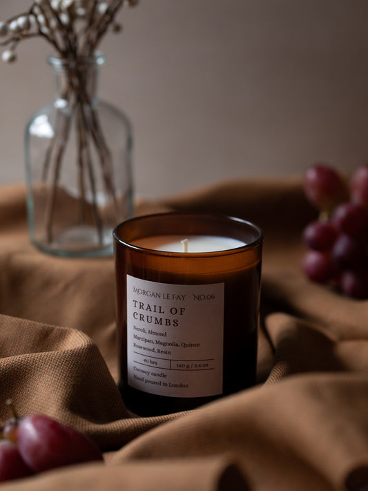 Candle labeled 'Trail of Crumbs' on a brown fabric surface with grapes and a vase in the background.